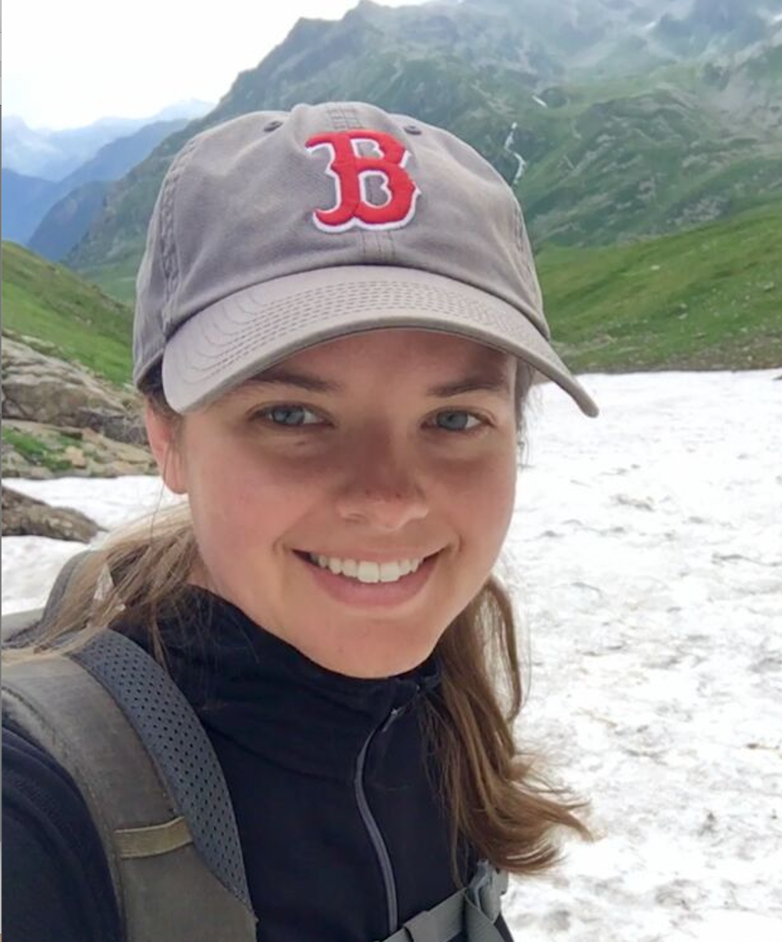 Close-up photograph of Shauna, a smiling young white woman wearing a Boston Red Sox ball cap and a hiking backpack, standing on a beach in front of steep green mountains
