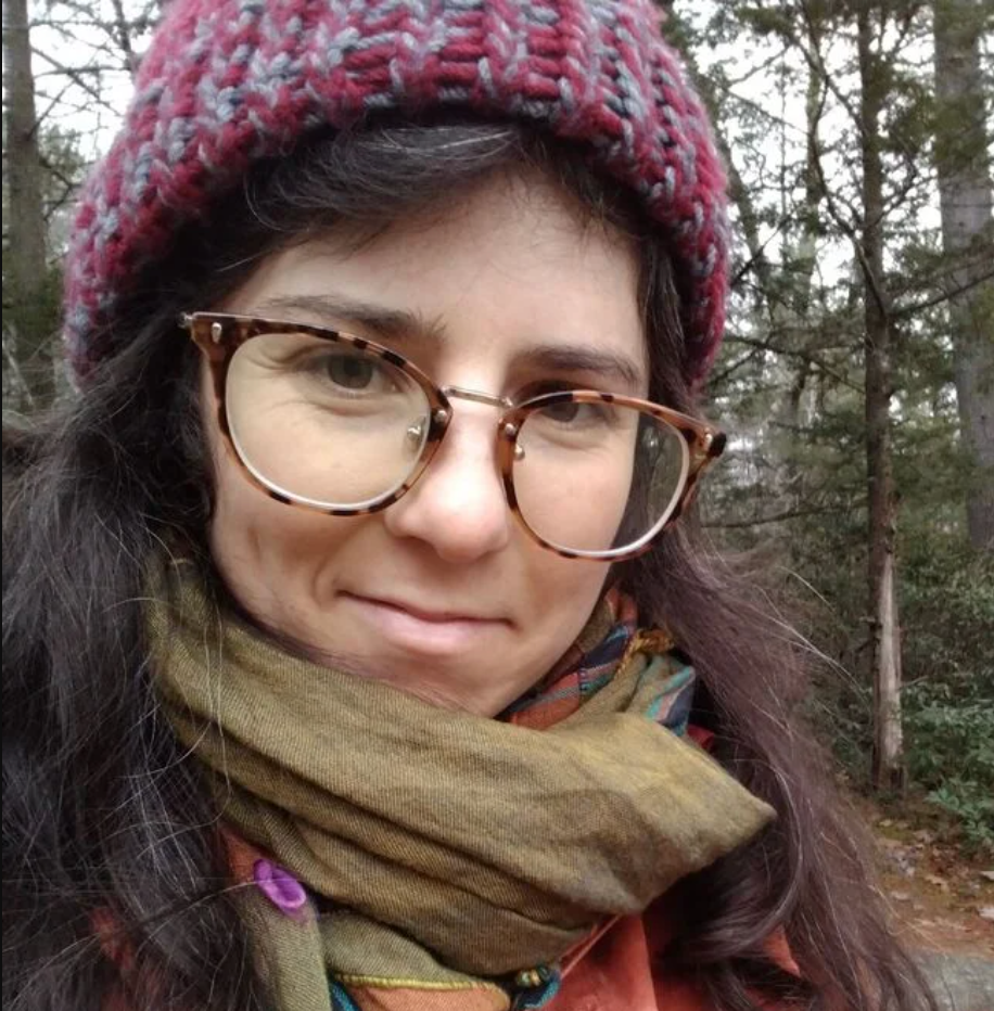 Close-up photograph of Clare, a white woman with dark hair, glasses, and a cozy winter scarf and hat, standing in a forest