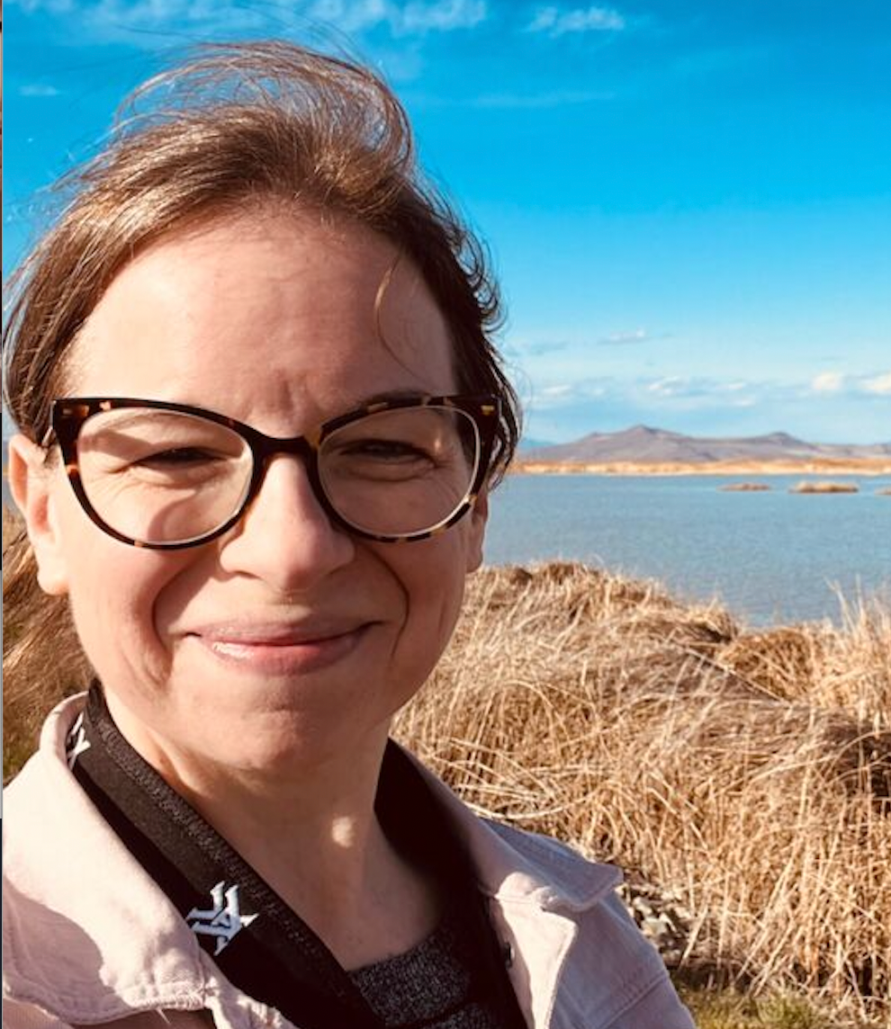 Close-up photograph of Claire, a smiling white woman with light brown hair and glasses standing on a grassy cliff overlooking the ocean on a sunny, windy day