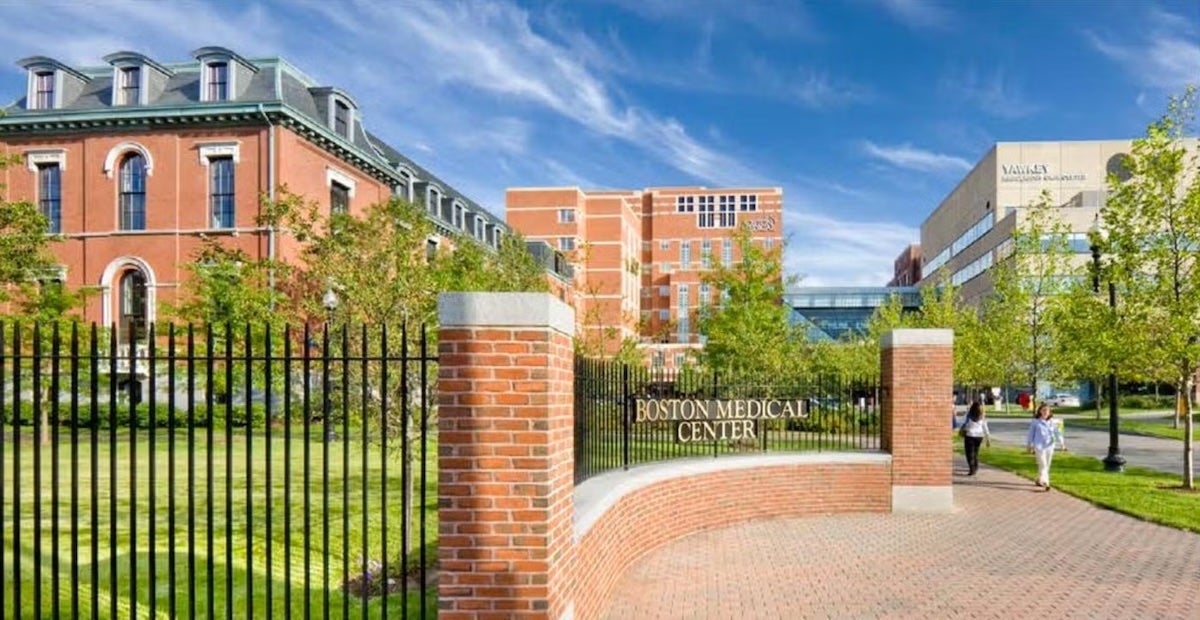 Photo of the Boston University Medical Campus. A bricked driveway curves in front of a tall iron fence enclosing a grassy lawn, with brick and concrete buildings visible on the far side