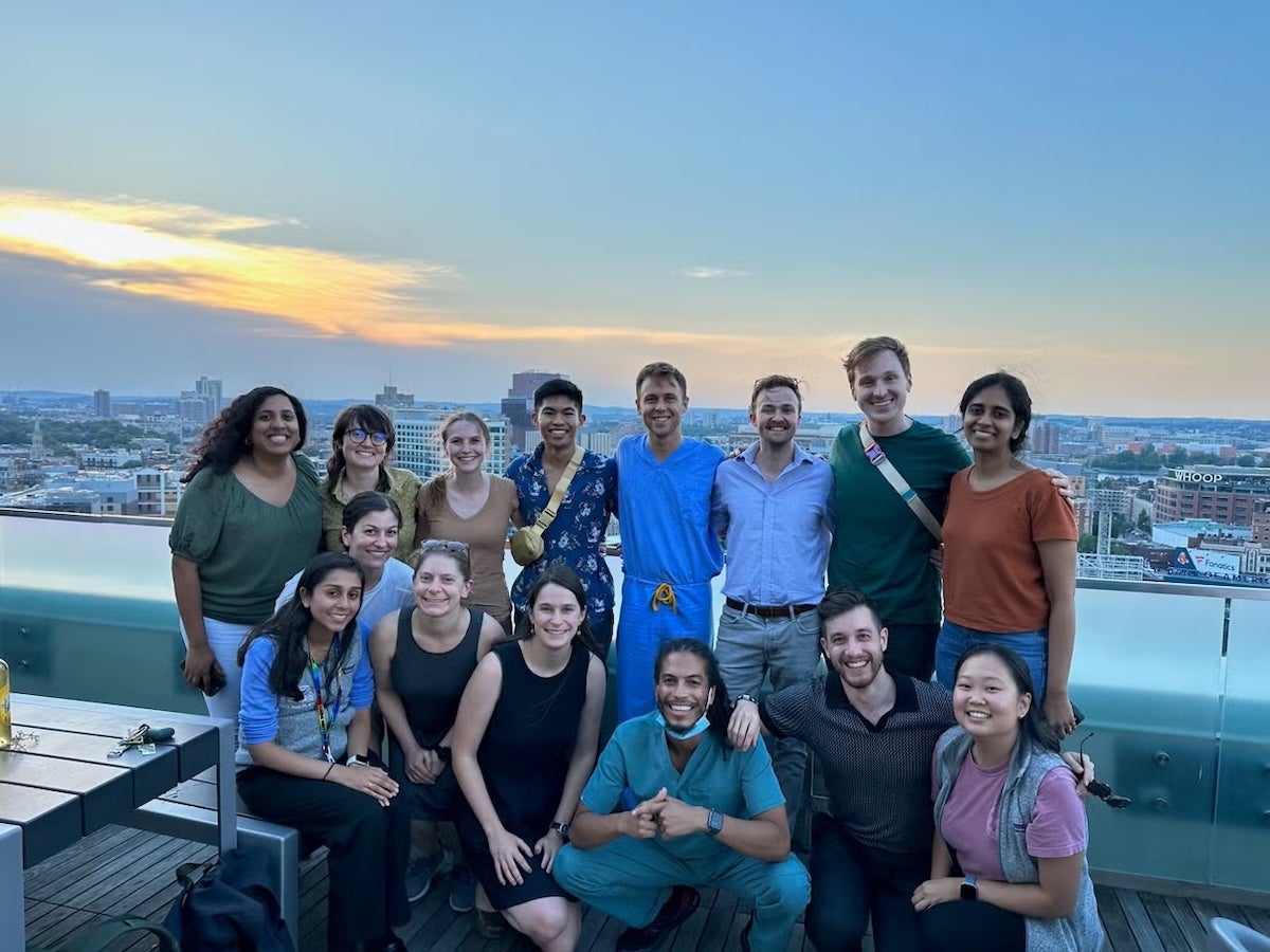 Group photo: members of HER Team 2023–2024 (fifteen individuals) on a BMC roof deck that overlooks the Boston skyline