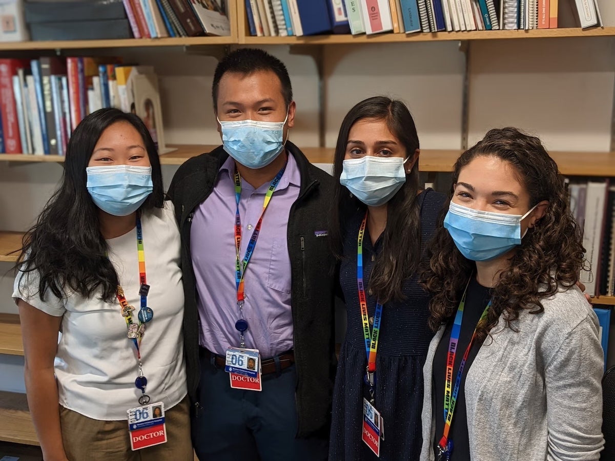 Group photo: four individuals wearing surgical masks and lanyards, standing in front of a book case