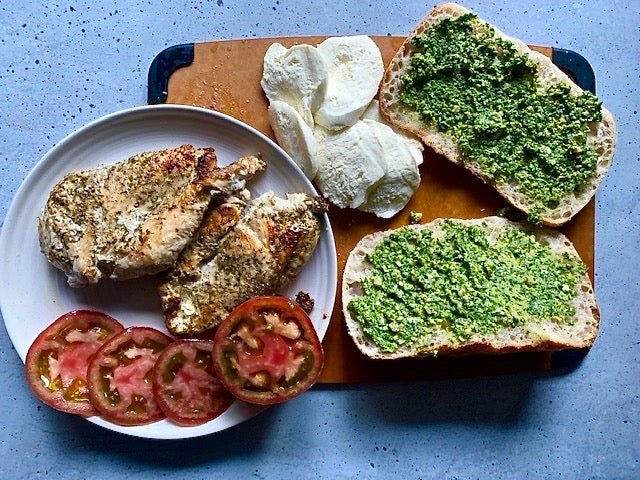 Display of cooked chicken breast, sliced tomatoes, sliced mozzarella, and bread with pesto spread on it. Displayed on a cutting board and plate. 