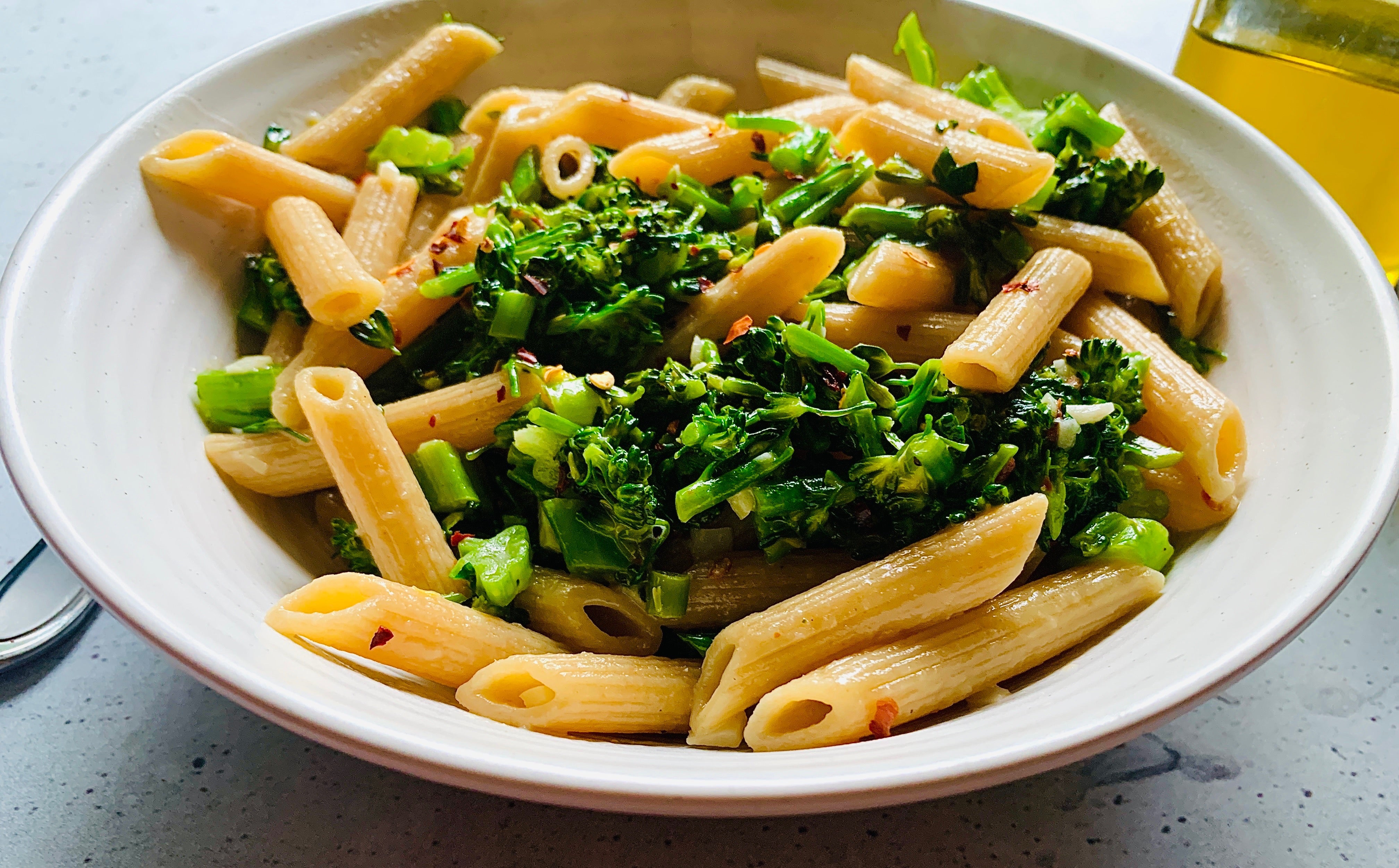Un tazón poco profundo lleno de penne de trigo integral, brócolini verde brillante picado, ajo y hojuelas de pimiento rojo.