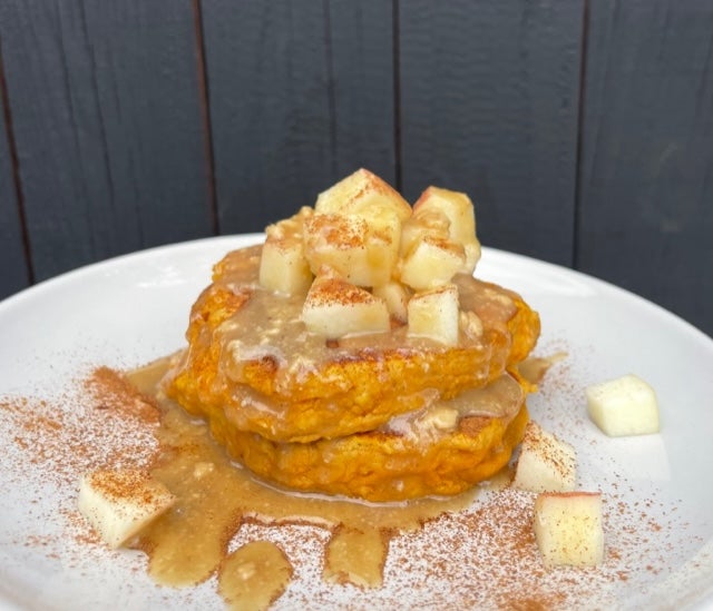 White plate stacked with the sweet potato pancaked, topped with diced apple and maple tahini sauce. Set against the bakground of a blue wall. 