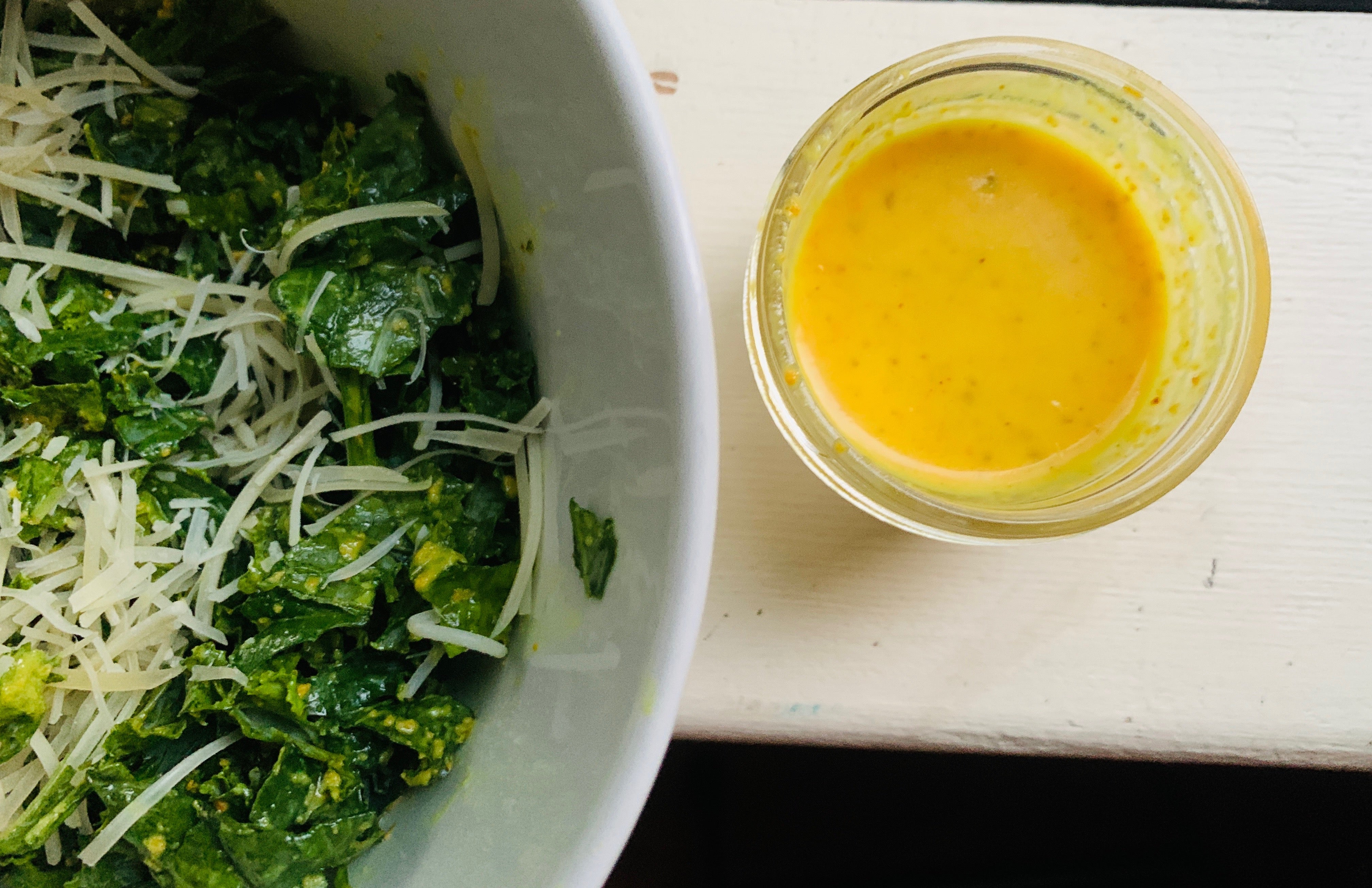 Turmeric Lemon Vinaigrette in a small glass jar, next to a large bowl of kale salad with parmesan cheese on top. 