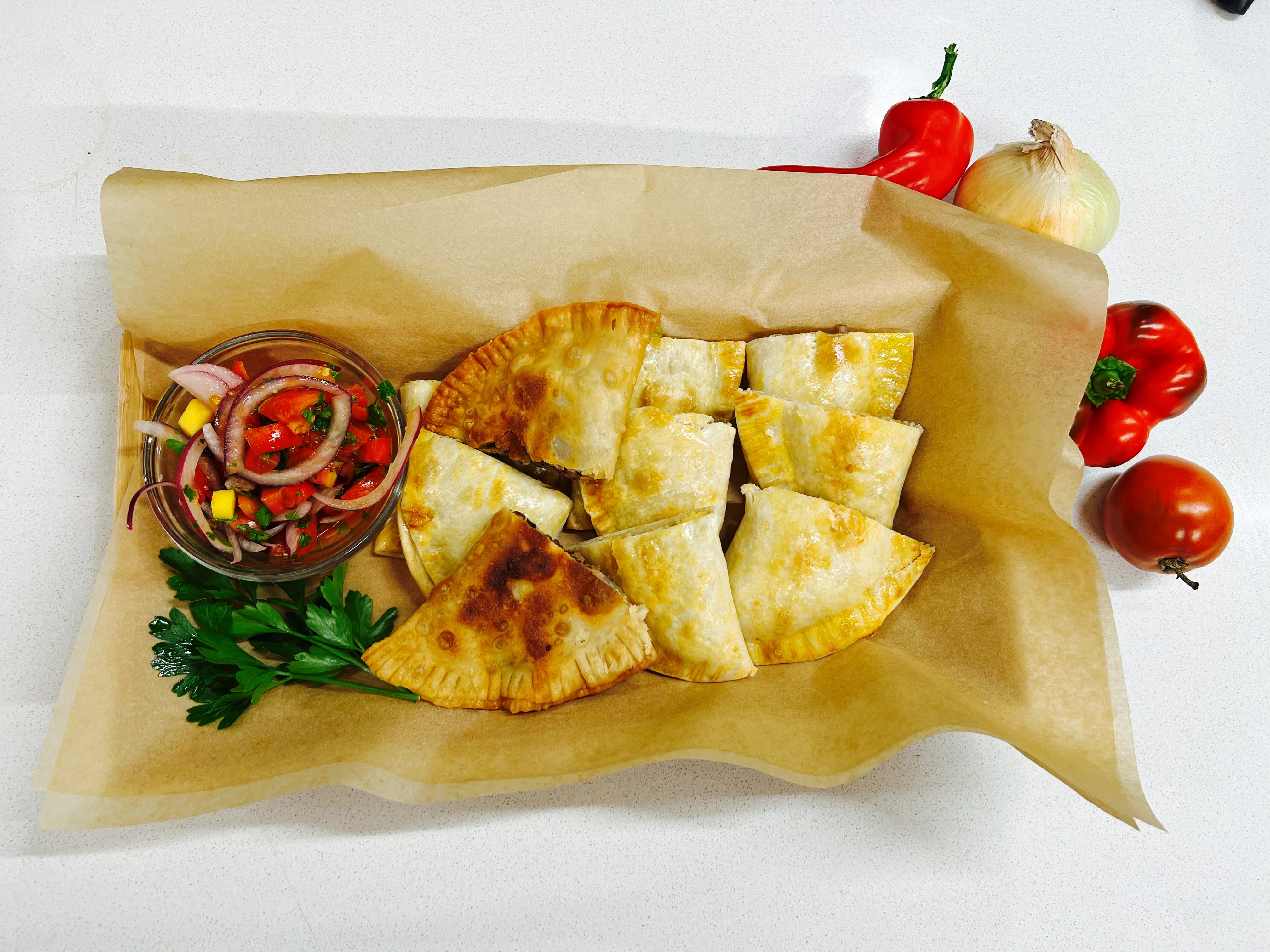 Baked empanadas with a bowl of fresh salsa on a platter lined with parchment paper. Tomatoes and parsley on the side as decoration.  