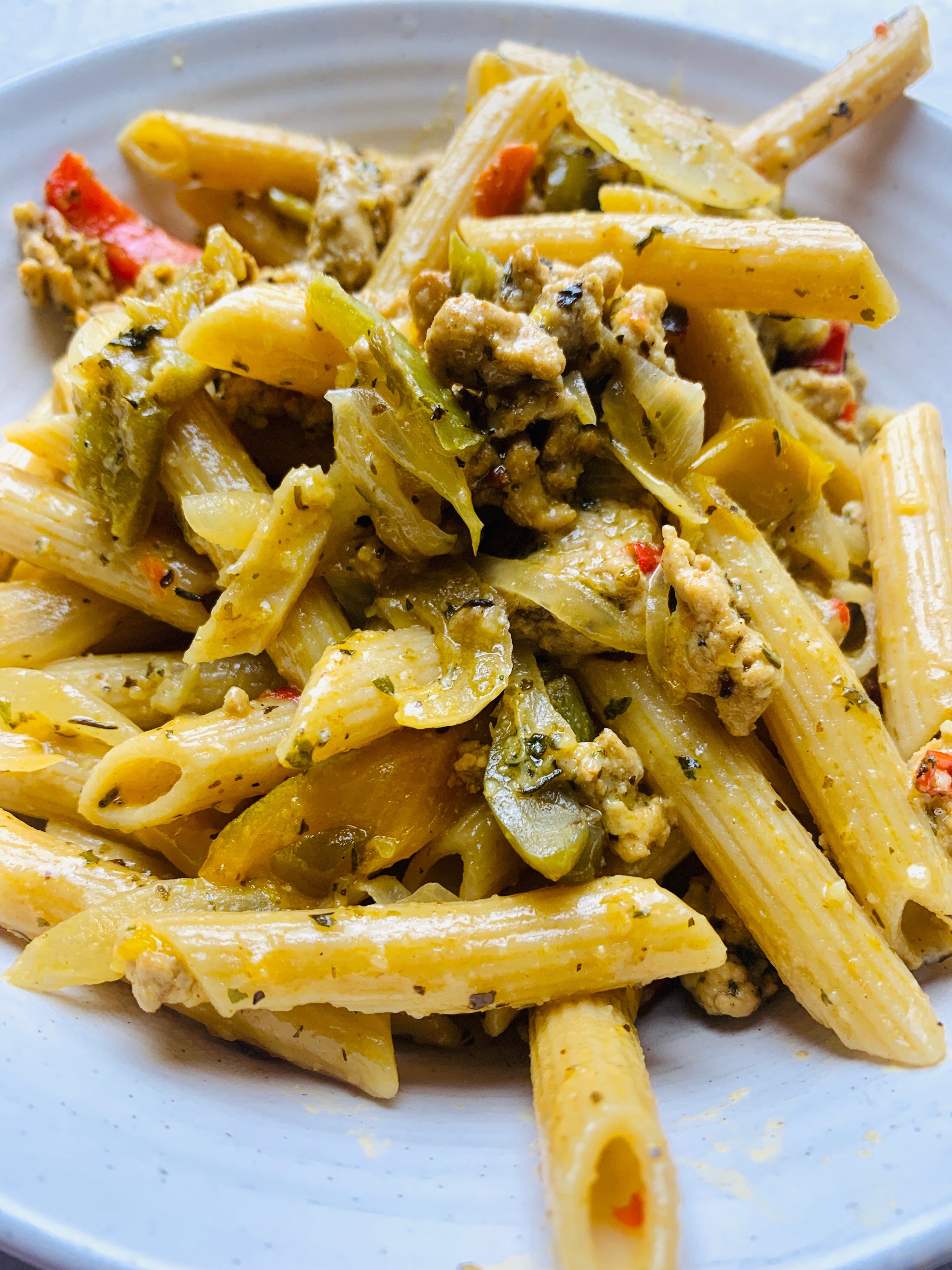 Penne pasta with sausage in a shallow white bowl, with herbs, pepeprs and onions