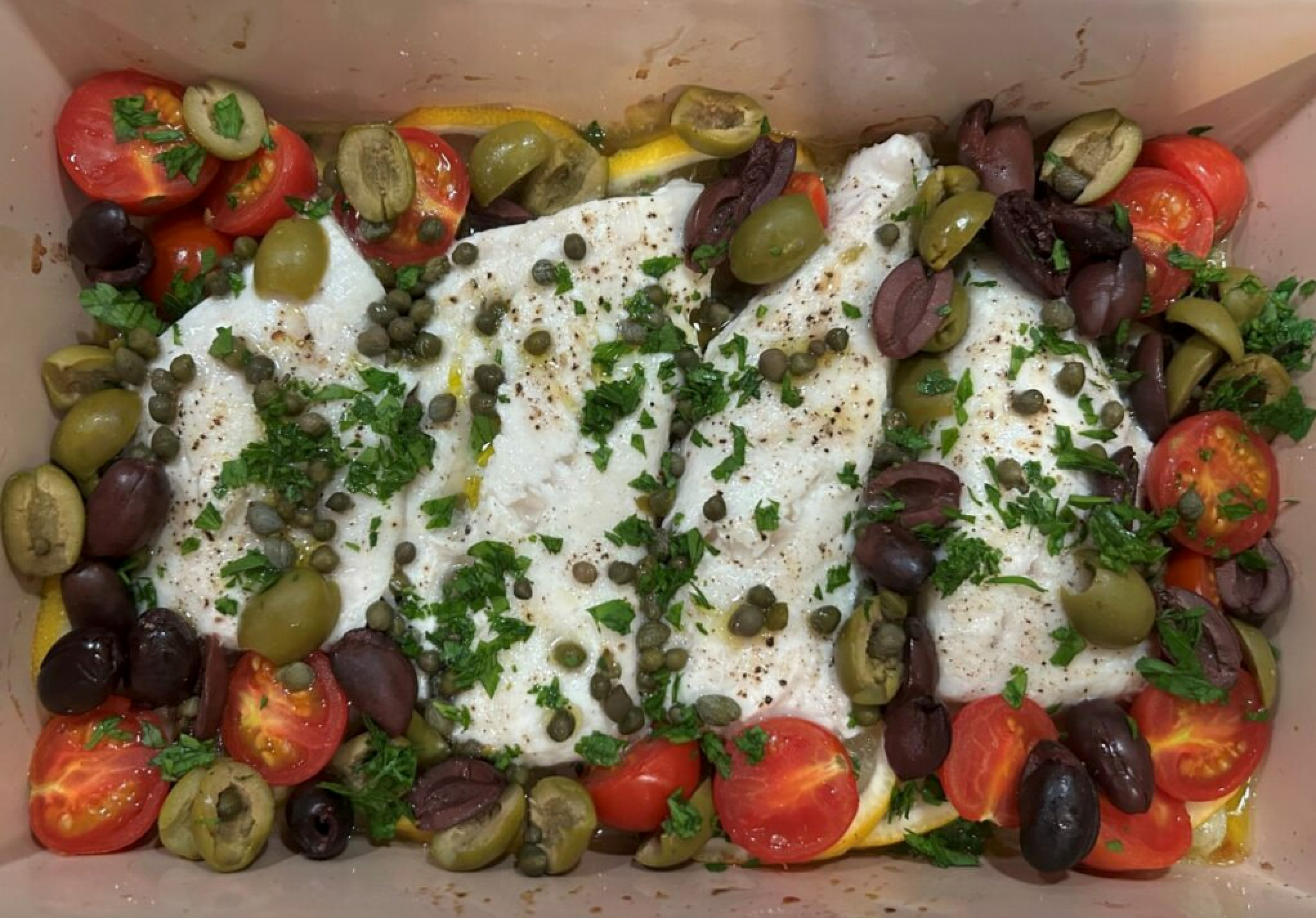 Baked white fish fillets topped with cherry tomatoes, olives, capers, parsley, and lemon slices.
