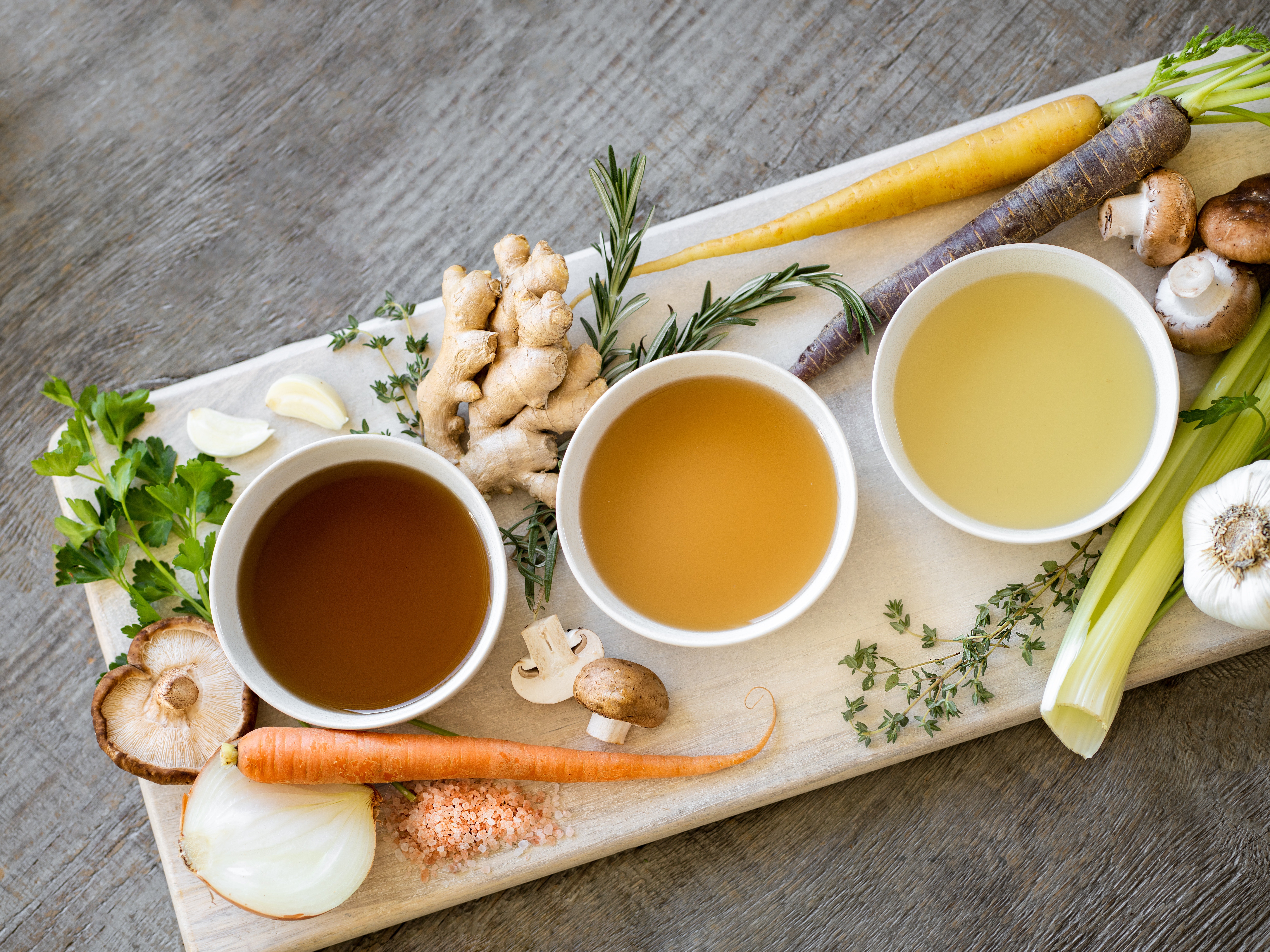 A wooden board covered with various vegetables (carrots, ginger, celery, mushrooms) and three small bowls filled with broth of different hues. 