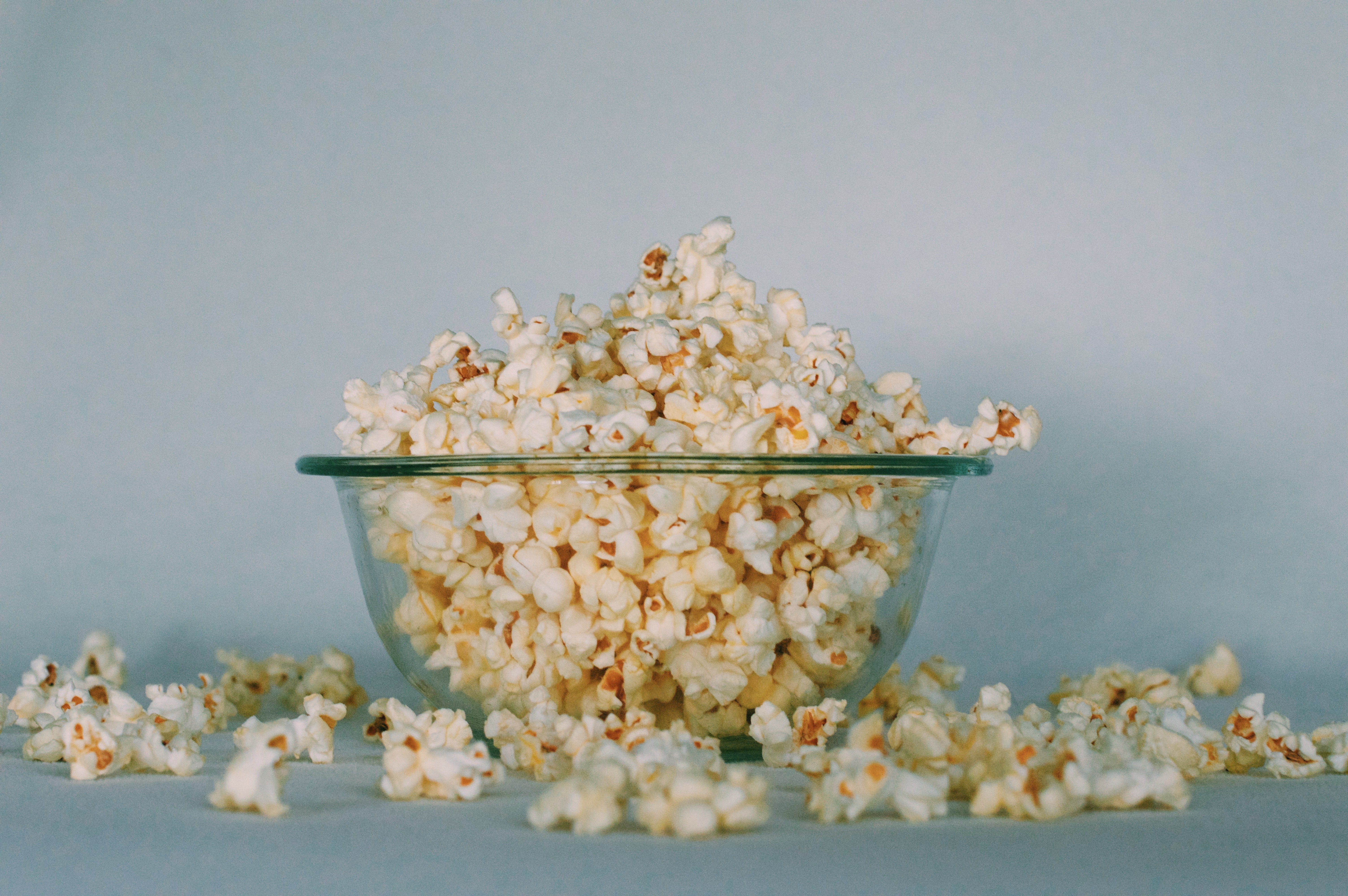 Glass bowl filled with popcorn. Popcorn overflowing onto the counter beneath the bowl. 