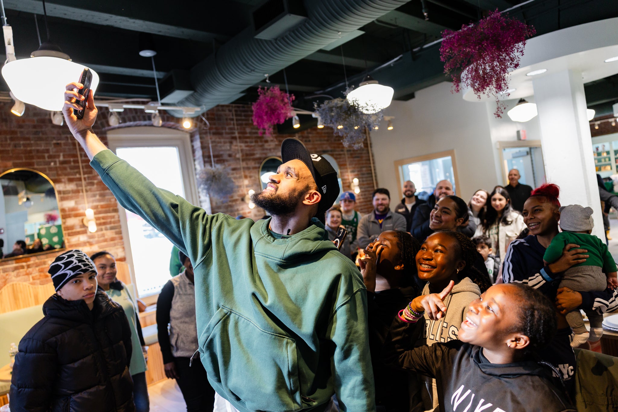A smilling and laughing group of mixed-aged people (mother holding a toddler, children and teens) in a restaurant gathers around a Boston Celtics player for a selfie. (Event photo from the fourth annual Curbside Care Celebration at the Little Cocoa Bean in the Boston Children’s Museum, hosted by the Celtics Shamrock Foundation with Tufts Health Plan.)