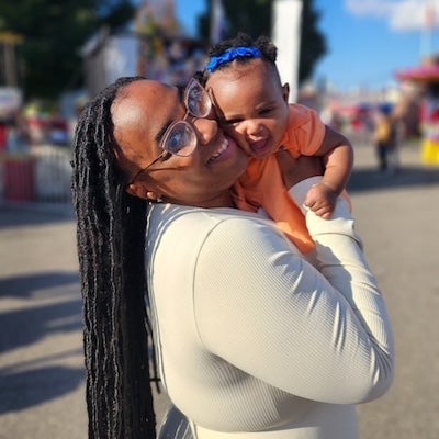 A smiling Kibriyaa holds Adaira up and smiling while standing on a sunny sidewalk
