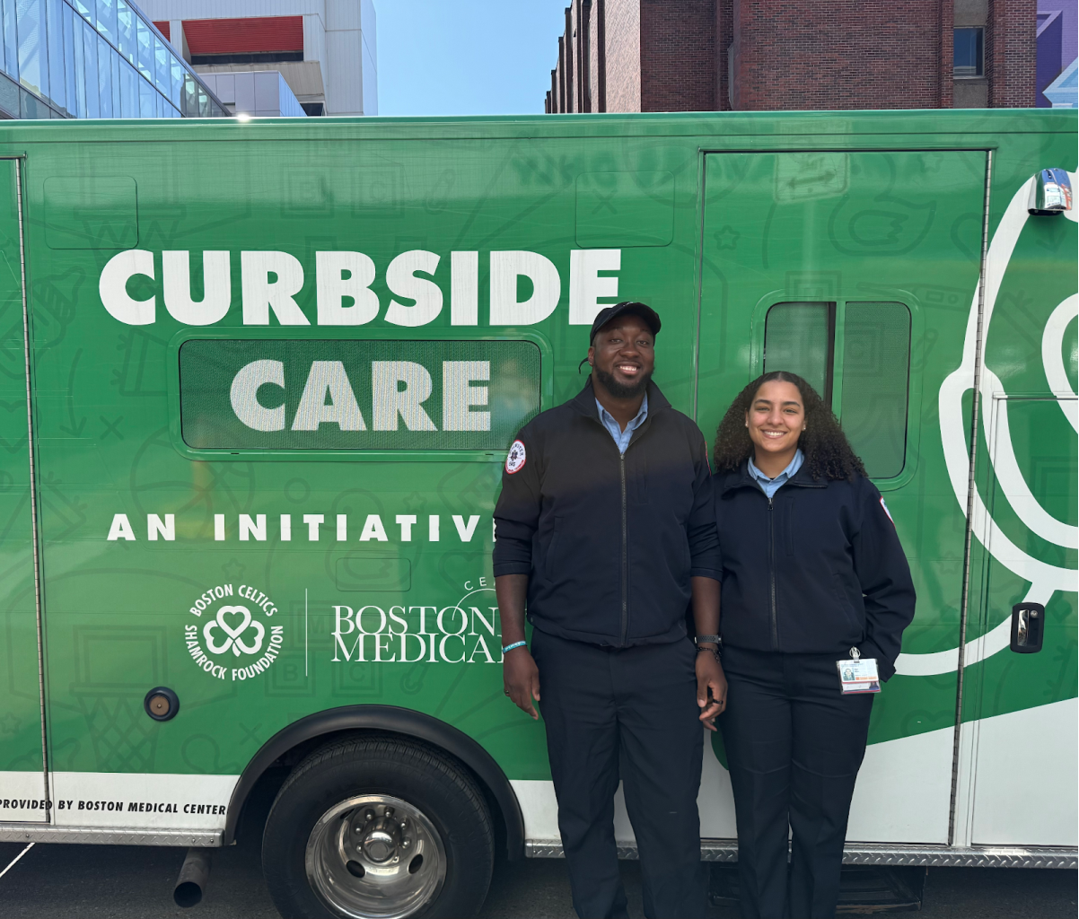 Two Curbside Care drivers wearing EMT uniforms smiling as they stand in front of the bright green BMC Curbside Care mobile medical services van