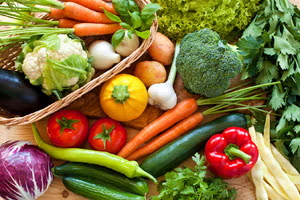Photograph of a basket overflowing with many colorful whole vegetables, such as tomatoes, cucumber, bell peppers, and garlic