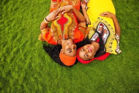 Two Black women wearing traditional African clothing lie on a grassy lawn and smile at the camera
