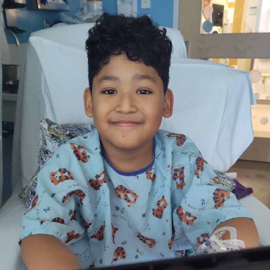 BMC pediatric surgery patient Dylan, sitting in a hospital bed and wearing a hospital gown with a cheerful print on it, smiles as he plays a game of Batman-themed Connect Four