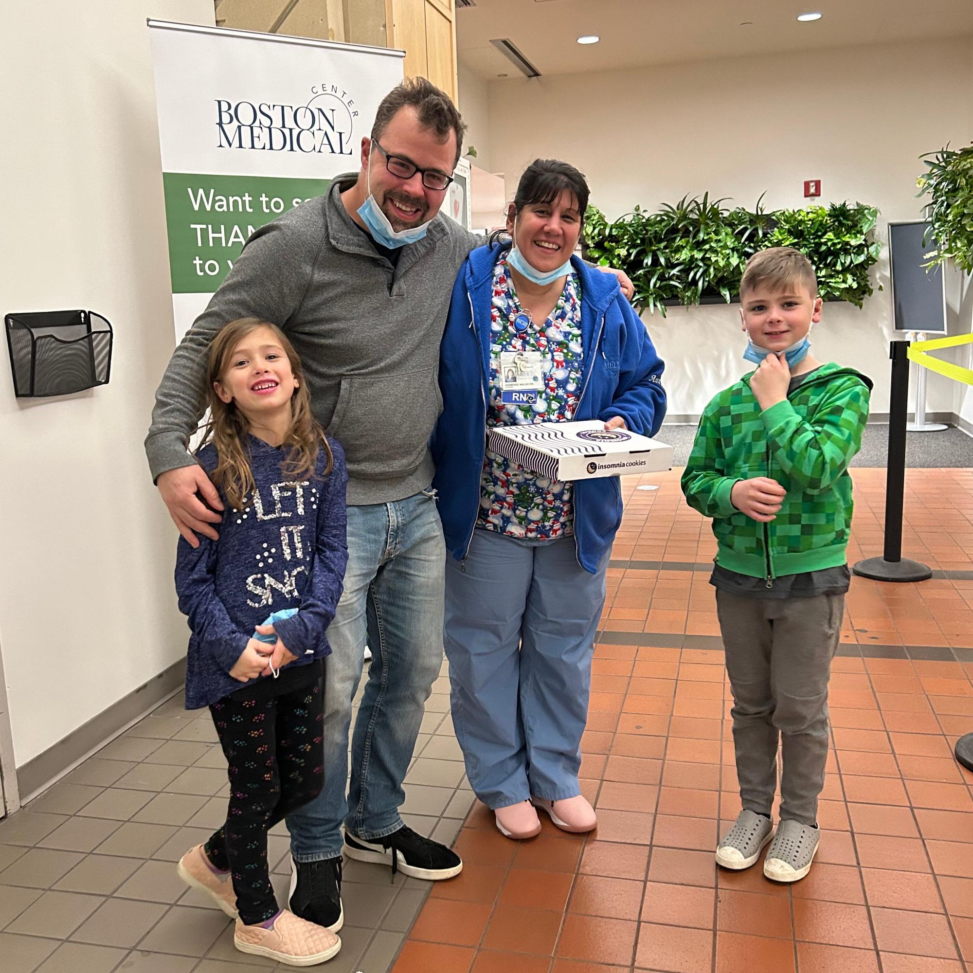 Former BMC patient Joe Tierney with his family, standing in the lobby of BMC with a box of treats they are gifting the pediatric nursing team in thanks for the lifesaving care he received in 1994