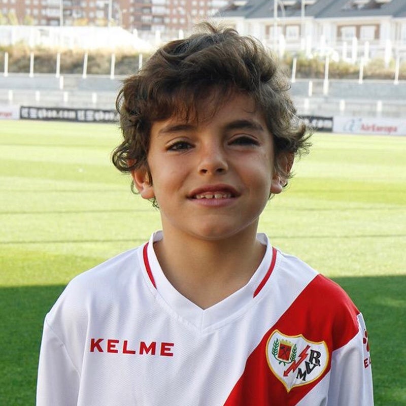 A child with brown hair wearing a red and white soccer jersey smiles while standing in a soccer field.