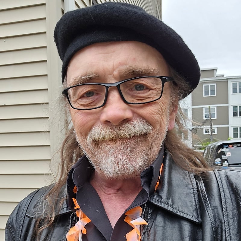 Headshot of BMC Cardiovascular Center patient Peter Goudy, a bearded white man wearing glasses, a black beret, black leather jacket, and Hawai'an lei necklace