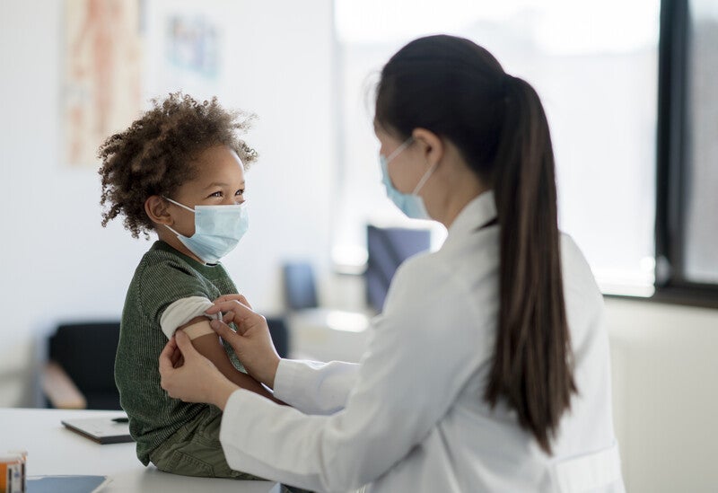A young child sits on an exam table and watches his female care provider place a bandage on his upper arm after administering a vaccine. The boy is dressed casually and both are wearing medical masks. (iStock)