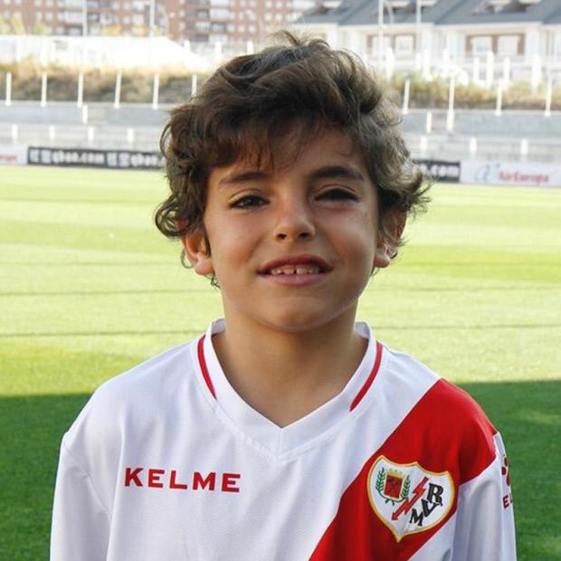 A child with brown hair wearing a red and white soccer jersey smiles while standing in a soccer field